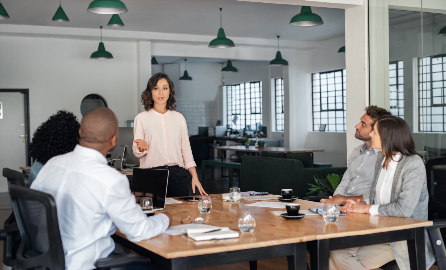 A woman stands presenting at the end of a conference table to two attentive colleagues, in a modern, well-lit office, representing a professional business meeting.