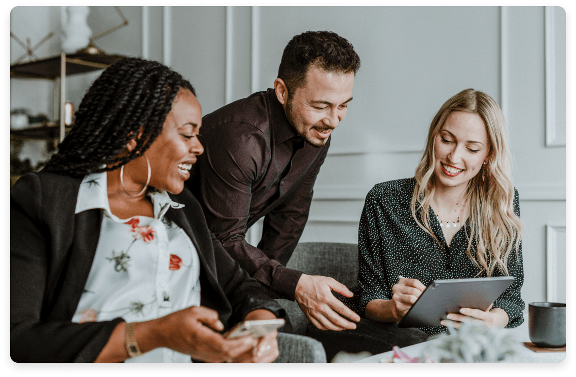 
                  Three cheerful colleagues collaborating with a digital tablet in an inviting office setting,
                  illustrating a team environment that values impact, learning, and strong connections.
                