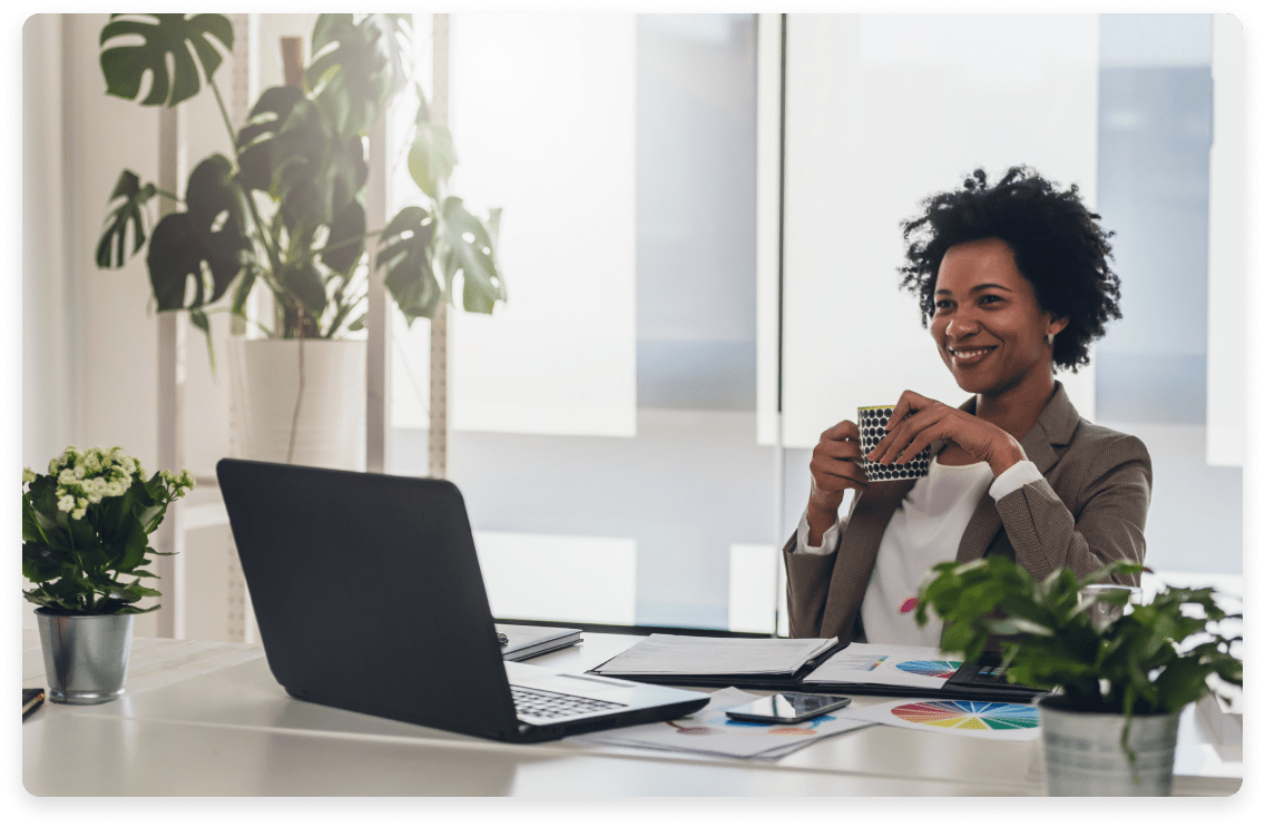 
                  A smiling professional woman with a coffee mug in hand sits at a well-organized desk with a laptop,
                  financial documents, and indoor plants, illustrating the efficient and pleasant environment of
                  fractional CFO services.
                