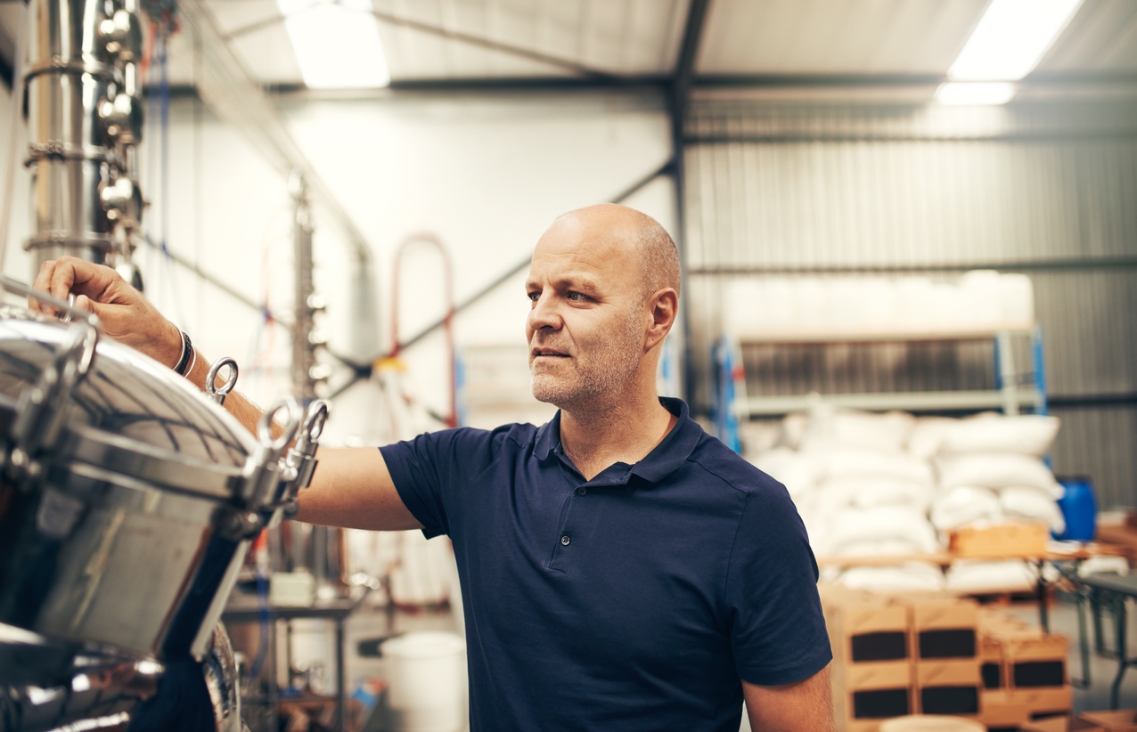 
                  A focused middle-aged man adjusting a valve on a shiny brewing tank in a distillery or brewery. He is wearing a dark polo shirt and is surrounded by industrial brewing equipment, with bags of raw materials stacked in the background, symbolizing hands-on quality control in beverage production.
                